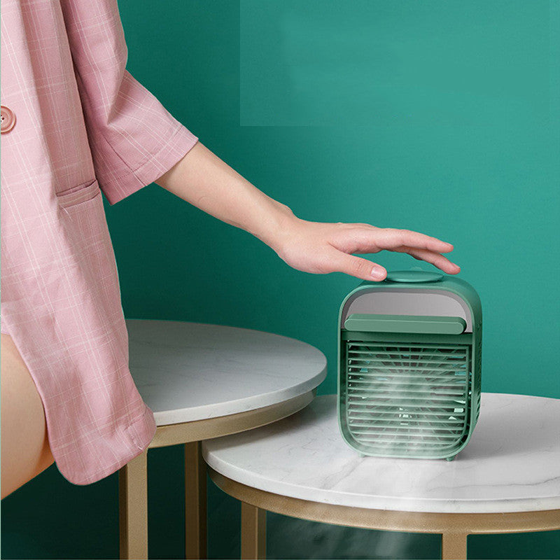 Desktop air conditioner fan with water spray on a table, being operated by a person in a pink outfit.